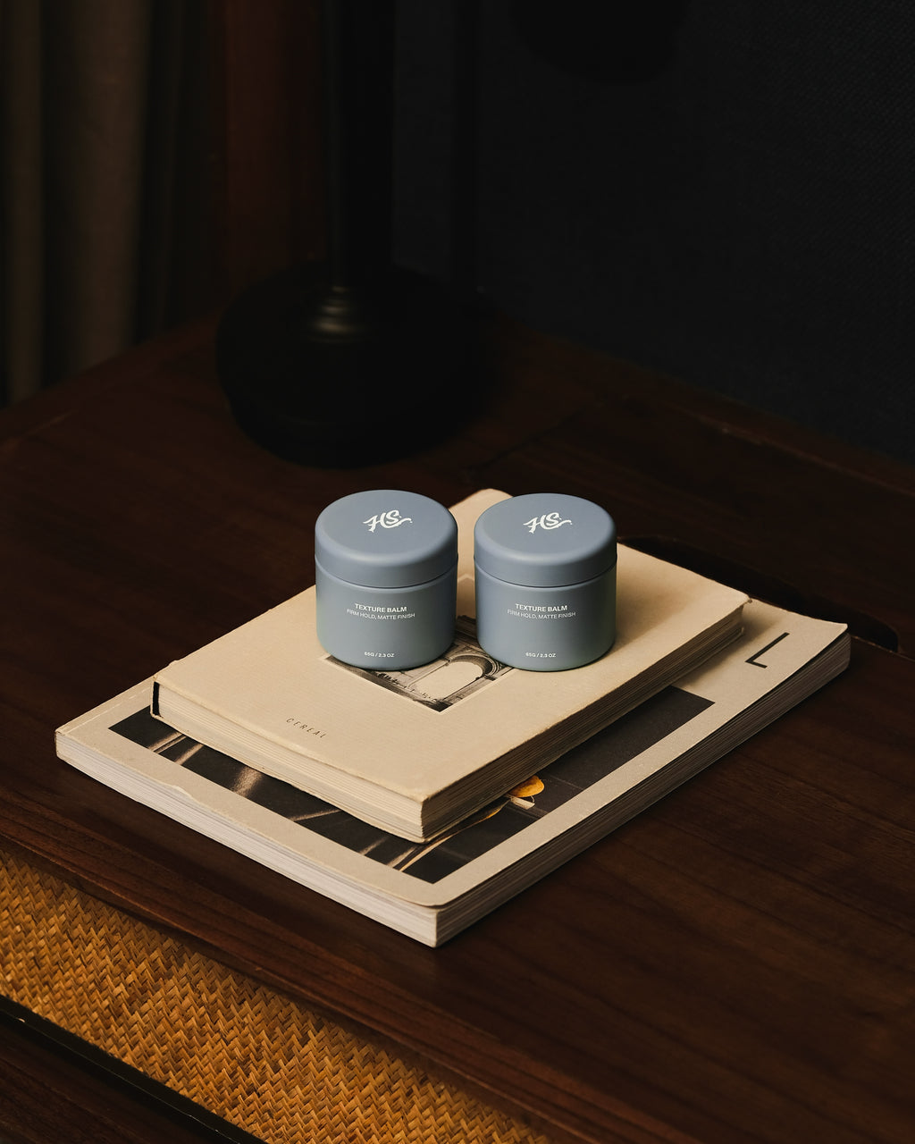 Two blue Hairsauce Texture Balm cylindrical jars on a book and wooden tray with a dark background