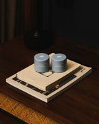 Two blue Hairsauce Texture Balm cylindrical jars on a book and wooden tray with a dark background