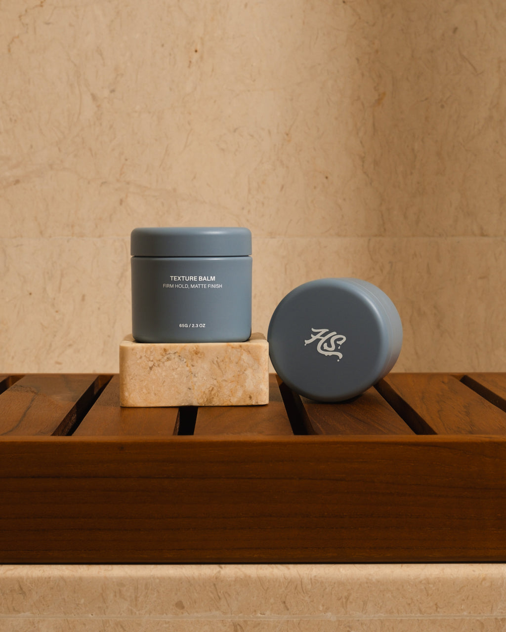 Two blue Hairsauce Texture Balm cylindrical jars on a wooden surface against a beige marble wall