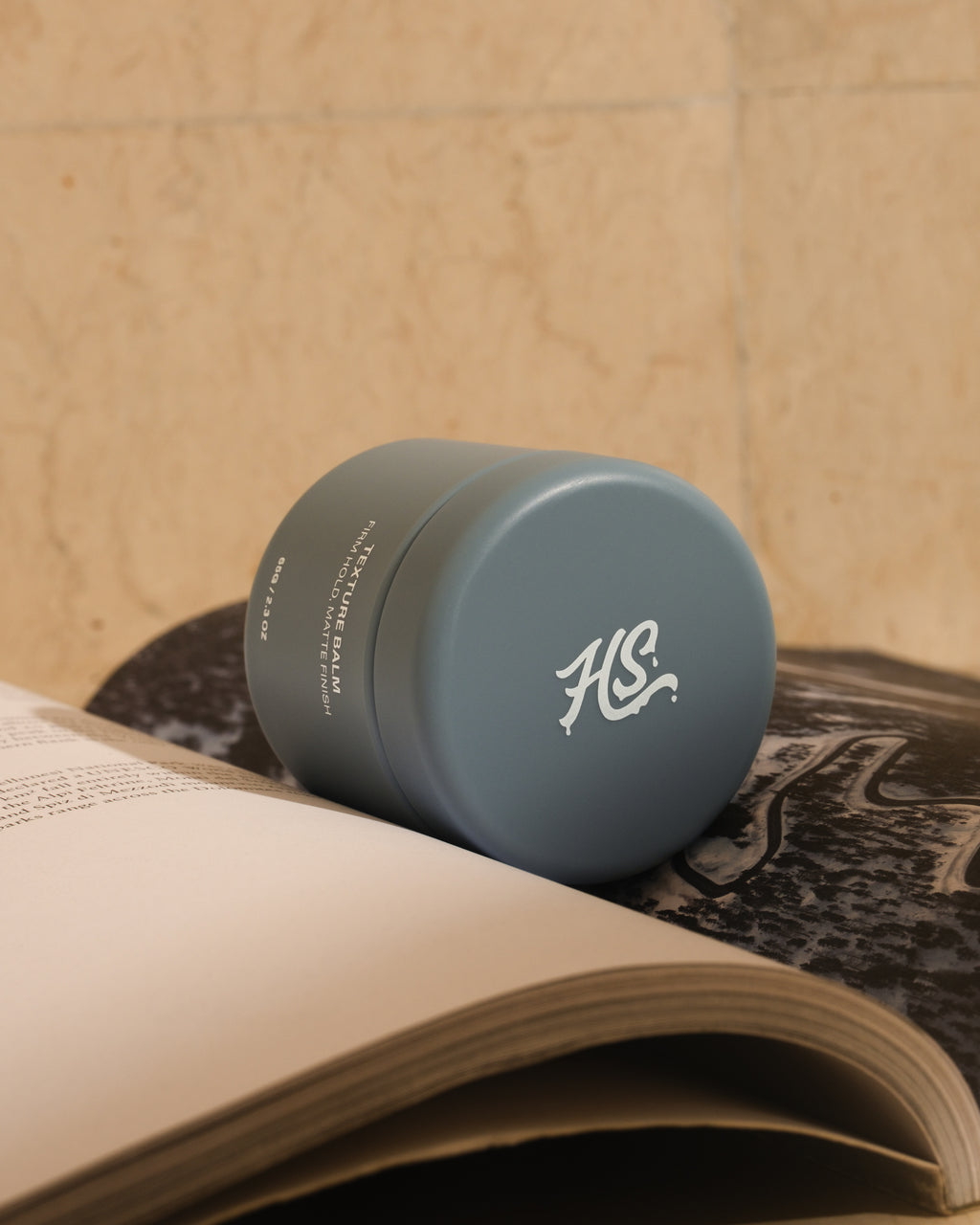 Blue Hairsauce Texture Balm cylindrical jar with a logo on top of an open book against a beige marble wall